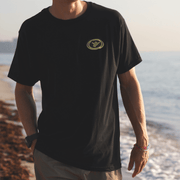 Person wearing a black t-shirt with a yellow Shaka logo on a beach