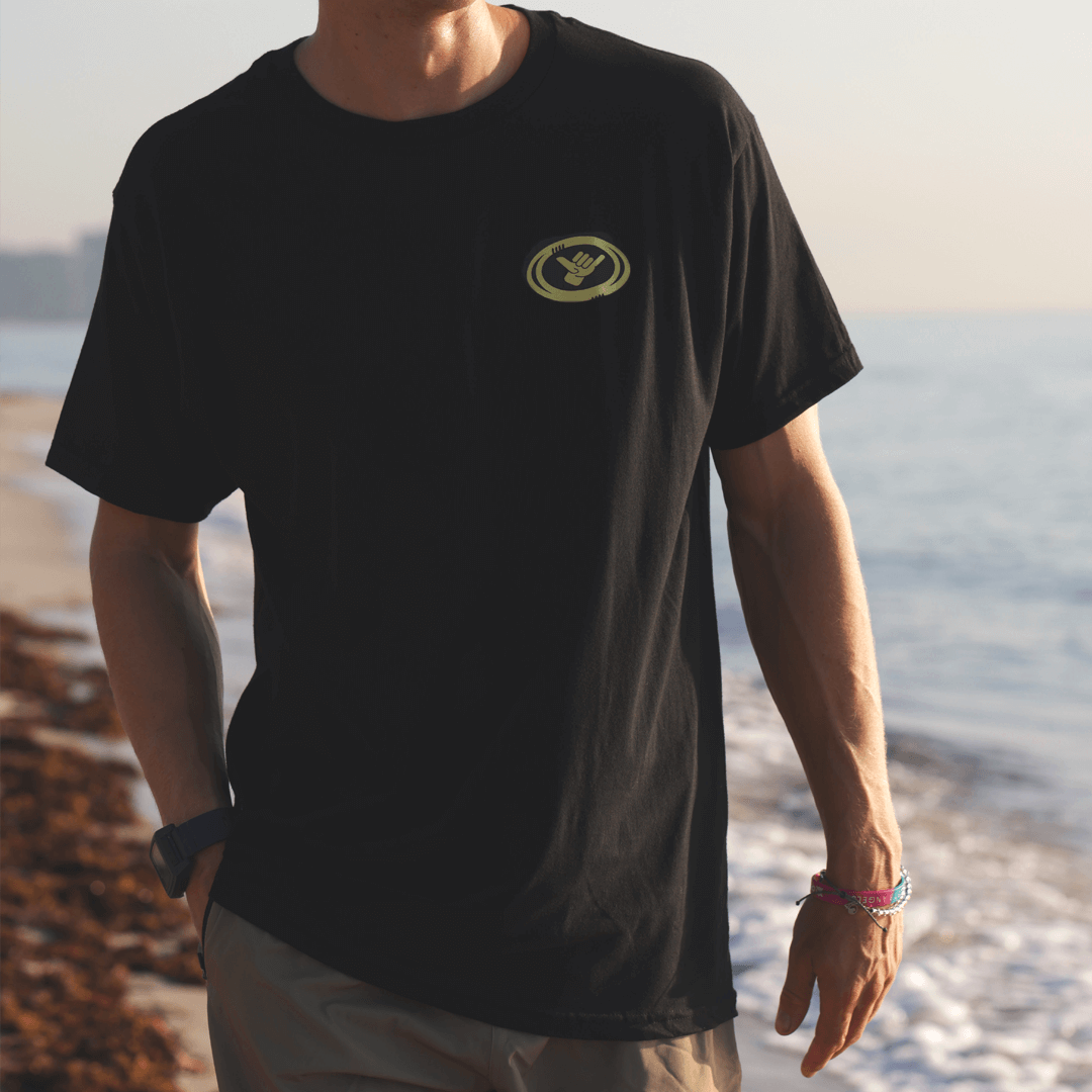 Person wearing a black t-shirt with a yellow Shaka logo on a beach