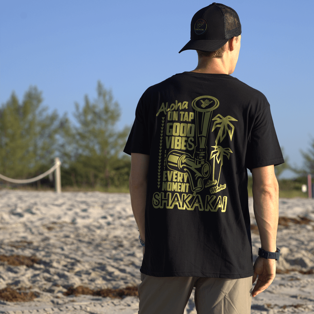 Person wearing a black t-shirt with yellow Shaka text and graphics on a beach