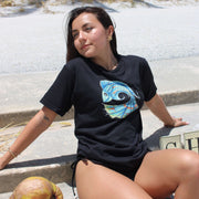 Woman wearing a black t-shirt with a colorful fish design on the front, sitting outdoors.