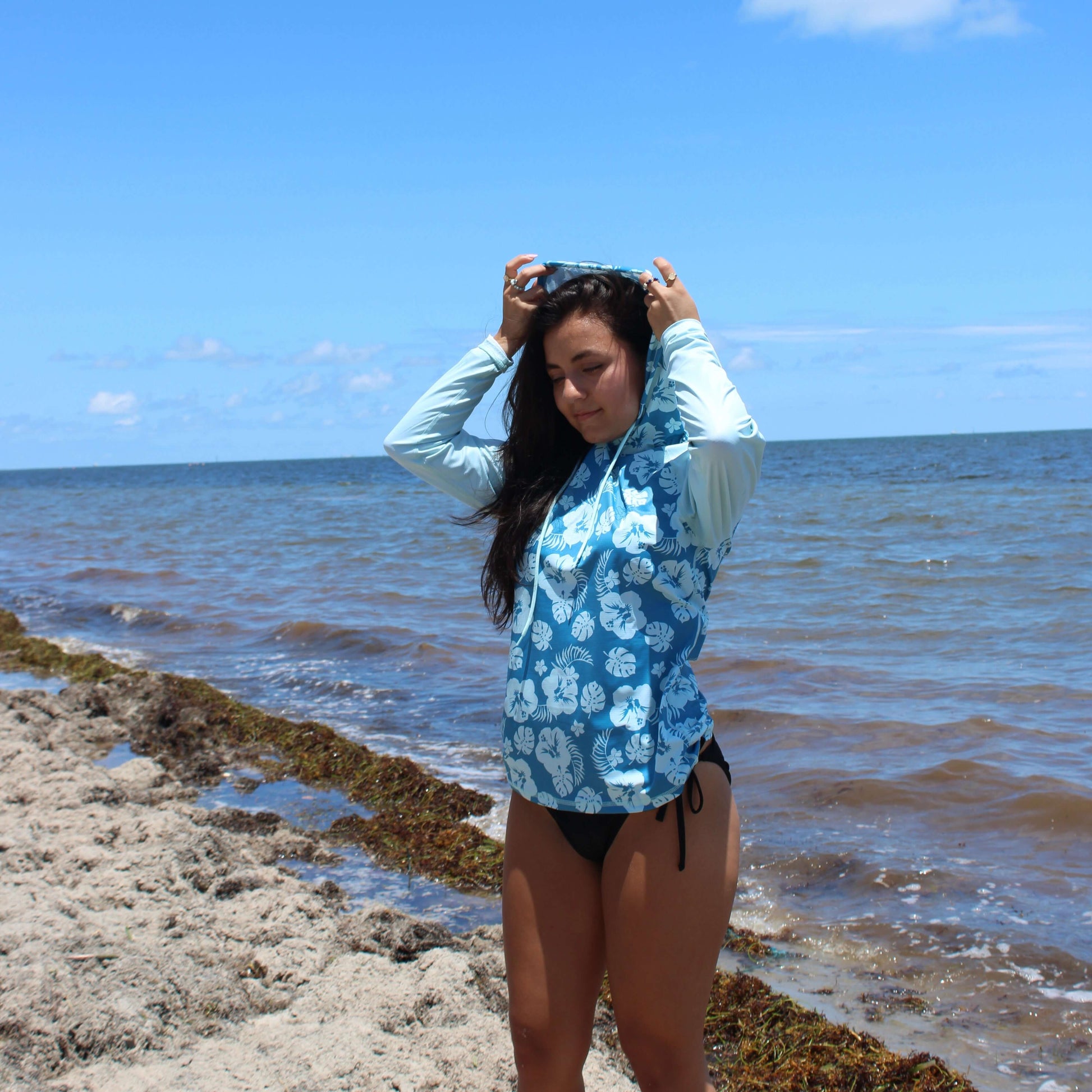 Woman wearing Plumeria LS Hooded Performance Shirt UPF 50 by the beach, enjoying sun and sea.