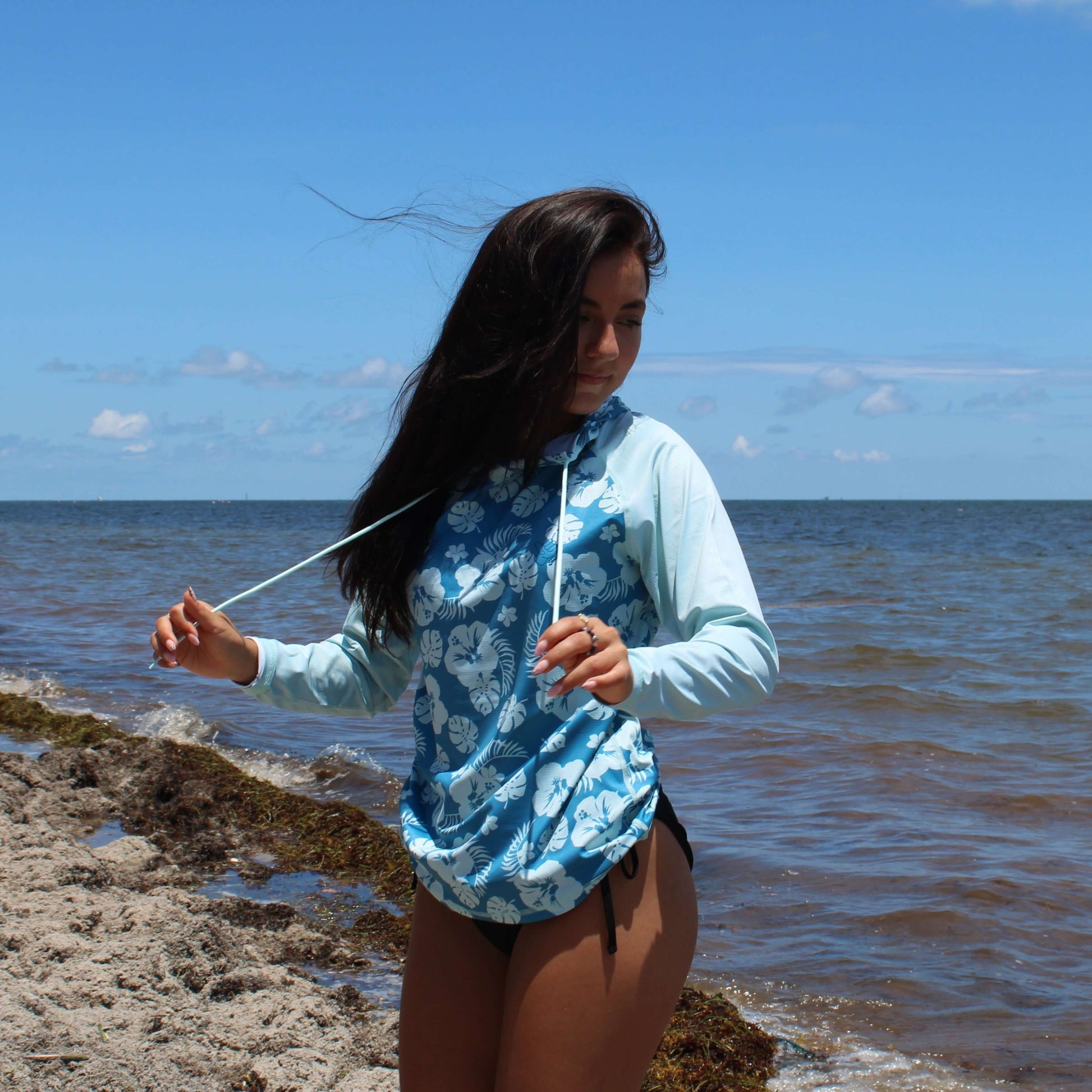 Woman wearing Plumeria LS Hooded Performance Shirt UPF 50 by the beach, enjoying sun protection with style.