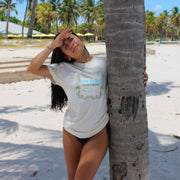 Woman wearing a white t shirt with tribal and Shaka graphic on the front by the beach