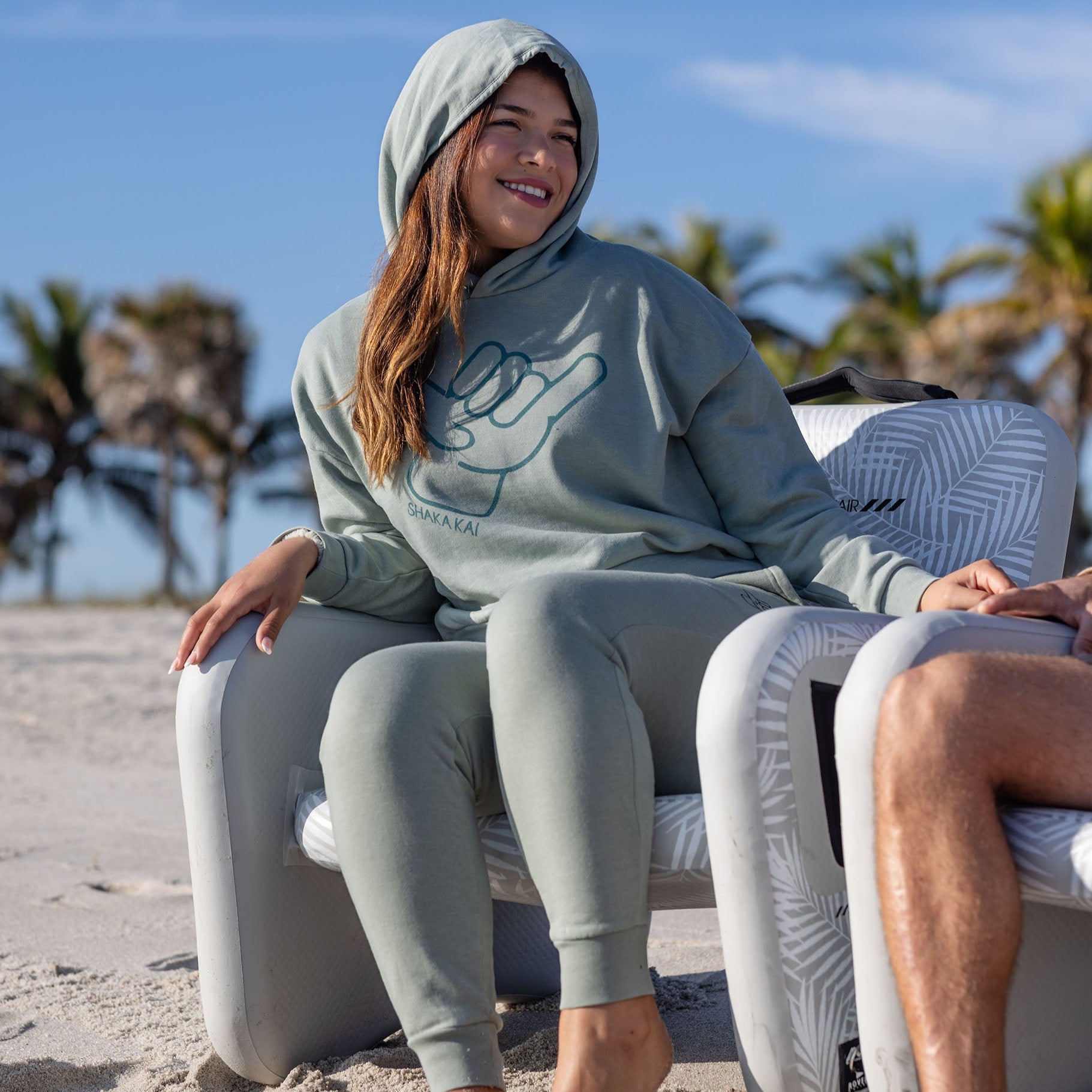 Model wearing Icon Womens Hoodie in a beach setting, showcasing casual comfort and island style.